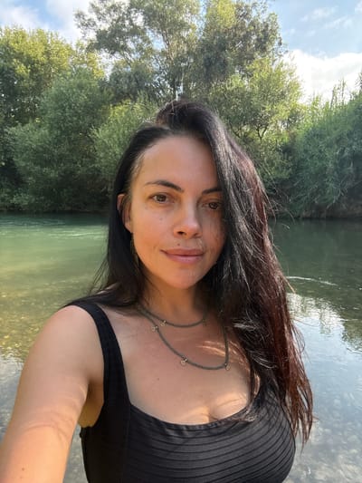In a selfie taken at the Jordan River, near the city of Mevoot HaHermon Regional Council, Israel, a woman with dark hair and brown eyes looks at the camera. She is wearing a black, ribbed tank top and a beaded necklace with small gold pendants. The river, with its clear, shallow water and rocky bottom, is visible in the foreground. Lush green trees and foliage line the riverbanks, and the sky above is partly cloudy. The sunlight casts dappled shadows on the water and on the woman's skin.