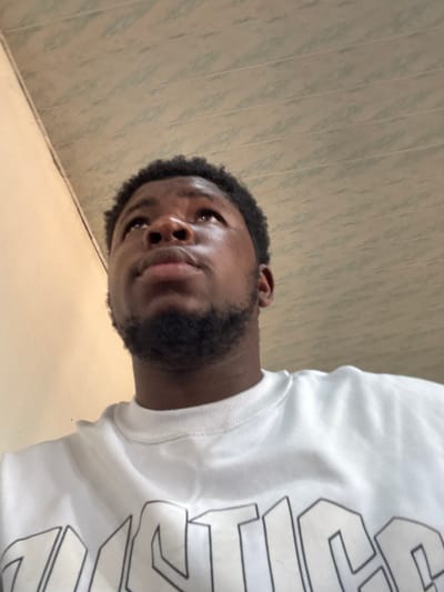 A low-angle image captures a male subject in Osu, Nigeria. The individual, an adult with dark skin, short dark curly hair, and a trimmed beard, occupies the lower portion of the frame, looking upwards and slightly to the viewer's right. He is dressed in a white long-sleeved crewneck sweatshirt featuring a partially visible black-outlined graphic print displaying the letters "USTICE". The background on the left consists of a plain, light-colored wall, while the upper right features a light-toned paneled ceiling exhibiting faint horizontal lines. No other subjects or specific objects are discernible.