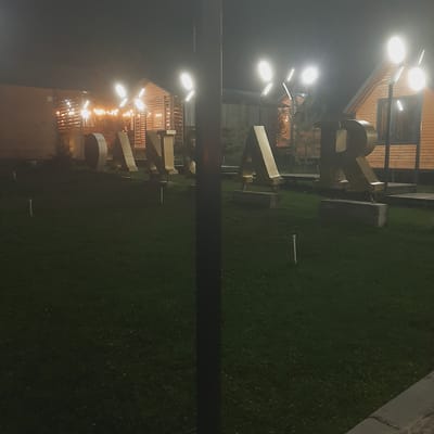 In Sosnovka, Kyrgyzstan, large, golden, three-dimensional letters spelling "ANSAR" are displayed on a well-maintained, dark green lawn at night. The letters are illuminated by bright, modern streetlights mounted on poles. Behind the letters, wooden buildings with pitched roofs and exterior lights are visible. The scene suggests an outdoor event space or a commercial establishment.