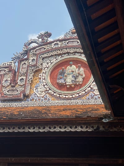 The image displays a highly ornate section of a traditional Vietnamese building's rooftop, set against a light blue sky with minimal cloud cover. The structure, located in Phú Xuân Ward, Hue City, Vietnam, features an extensively decorated gable. Central to this gable is a circular, terracotta-red medallion containing three relief figures. The central figure is a man wearing a tall, patterned hat and lighter-colored robes, flanked by two other men in darker, patterned robes, all standing front-facing.

Surrounding the central medallion and extending across the gable are intricate mosaic patterns composed of white, blue, and brown ceramic pieces, forming geometric designs and stylized floral motifs. Visible at the upper edges of the gable are sections of two stylized dragon sculptures. Below the elaborate gable, rows of weathered orange and reddish-brown terracotta roof tiles are present. To the right of the frame, a dark wooden architectural element, possibly an eave or part of an adjacent roof, diagonally descends into view. The scene is illuminated by bright daylight.