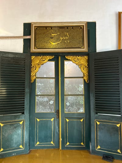A pair of dark green double doors with ornate gold trim are open, revealing louvered shutters on each side. A framed piece of Arabic calligraphy in gold lettering hangs above the doors. The doors have decorative glass panels with a floral pattern and brass door handles. The floor is covered in yellow tiles. The image appears to be taken inside a building, possibly a mosque or historical site, in the city of Cirebon, Indonesia.