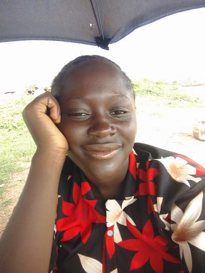 A medium shot captures a young person, likely a child or adolescent, from the chest up, posing for a selfie. The subject's face is turned slightly towards the camera, with their left arm bent and hand resting against their cheek, creating a relaxed and thoughtful pose. They are wearing a black shirt adorned with a bold floral pattern featuring red and off-white flowers with brown centers. The background is a bright, washed-out outdoor scene, suggesting it is daytime. To the left, there is a patch of green grass and what appears to be the edge of a dirt path. In the upper portion of the image, a dark fabric, possibly an umbrella or canopy, provides shade. To the right of the subject, in the distance, there are hints of more foliage and what looks like a light-colored cylindrical object. The overall impression is of a casual moment captured outdoors. This image appears to be taken in Gosa, Nigeria, a location known for its rural and semi-urban settings.