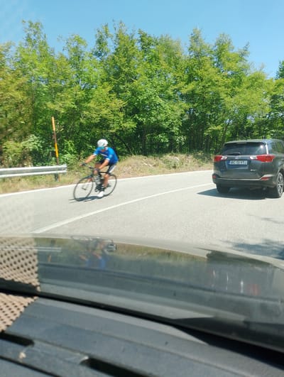 An exterior medium shot captures a sunny day scene on a road bordered by trees. In the foreground, the interior of a car is visible, with a dashboard and windshield reflecting the outside environment. The road itself is asphalt, with white lane markings. A lone cyclist, wearing a blue jersey, white helmet, and sunglasses, pedals a road bike along the right side of the road, their body blurred due to motion. Further down the road, a dark-colored SUV is parked on the right shoulder, its license plate reading "BC 13 YTJ." The surrounding environment is lush green with dense trees and foliage under a clear blue sky. The scene is set in Leonessa, Italy, indicated by the license plate format.