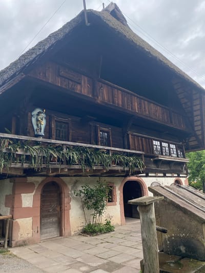 This image shows a historic Black Forest house in Gutach (Schwarzwaldbahn), Germany. The building is characterized by its traditional thatched roof and dark wooden facade. It has multiple stories with windows and a balcony decorated with plants. An ornamental figure adorns the facade below the balcony. Adjacent to the house is a stone structure, possibly a well or fountain, with water visibly dripping. The ground is paved with stone tiles. The scene appears to be a daytime exterior shot.