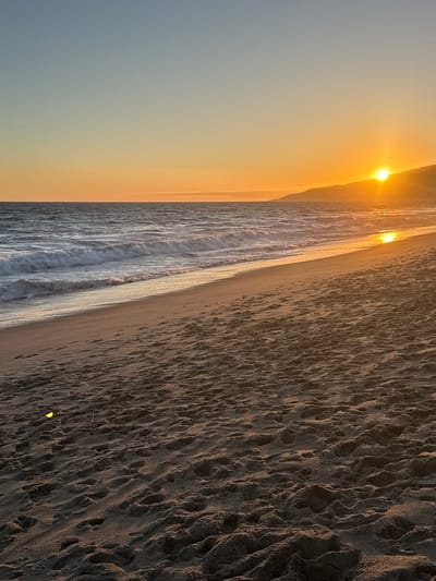 An ocean sunset scene at a beach in Malibu, California. The sun is low on the horizon, casting a warm orange glow across the sky and reflecting on the water. Gentle waves are rolling onto a sandy shore. The sand in the foreground is textured with footprints and appears to have some small debris on it. In the distance, a hilly coastline is silhouetted against the sunset. The sky transitions from a light blue at the top to a warm orange near the horizon.