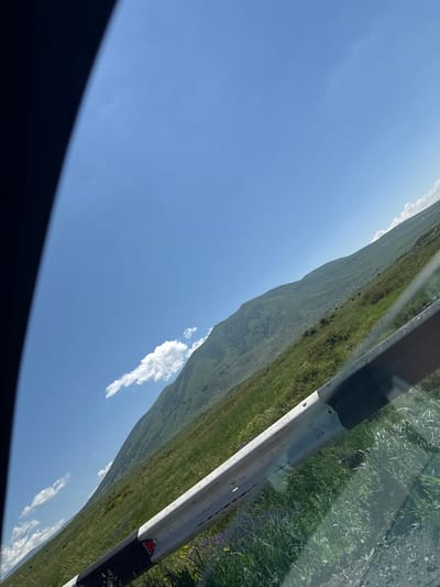 An outdoor, eye-level, medium shot captures a mountainous landscape in Saghmosavan, Armenia. The sky is a clear, bright blue with a few scattered white clouds. The foreground is dominated by a guardrail, its white and black stripes slightly weathered. Beyond the guardrail, a vast, green hillside slopes upwards, dotted with patches of grass and wildflowers. The hillside is partly obscured by a shadow in the upper left corner of the image. The overall scene is serene and natural, with no people or immediate activity depicted.