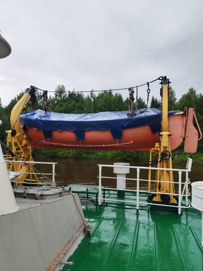 An orange lifeboat is suspended by a yellow davit on a green-decked ship. The lifeboat is covered with a blue tarp. The ship is docked near a tree-lined riverbank. The sky is overcast. This scene is located in the city of Vytegra, Russia, on the Vytegra River.