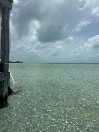 The image displays an outdoor scene depicting a calm, shallow body of water in San Pedro Town, Belize, viewed from a wooden structure. The lower two-thirds of the frame are occupied by clear, turquoise-green water, characterized by surface ripples that create intricate, shimmering light patterns on the visible seabed. On the far left, a weathered wooden support structure, consisting of vertical posts and horizontal planks, is partially visible. A white fabric panel hangs from this structure, descending towards the water and making contact with it at the bottom left. A flat horizon line extends across the image, separating the water from the sky. In the mid-ground, a low-lying landmass densely covered with green vegetation is visible on the left side of the horizon. Several small, dark, vertical markers are sparsely distributed across the distant water surface. The upper third of the image is dominated by an overcast sky, featuring extensive grey clouds with intermittent patches of lighter blue sky, particularly towards the right. No human or animal subjects are present in the frame.