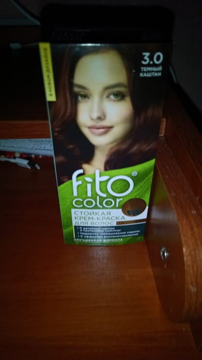 The image shows a box of Fito Color hair dye, shade 3.0, described as "Dark Chestnut." The box features a portrait of a woman with brown hair and green eyes. The text on the box is in Russian and highlights features like " стойкая крем-краска для волос" (lasting cream-dye for hair), "с целебным маслом и протеинами пшеницы" (with healing oil and wheat proteins), "надежное закрашивание седины" (reliable gray coverage), "с эффектом биоламинирования" (with bio-lamination effect), and "улучшенная формула" (improved formula). The box is placed on a wooden surface, possibly a table or shelf. The background is dark and out of focus. There is no discernible indication of the city of Balkhash, Kazakhstan, or any other specific location. The image appears to be a product photo taken indoors with artificial lighting.