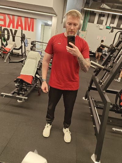 An image captured in a gym located in Nahariya, Israel, shows a male subject taking a mirror selfie. The male, positioned centrally, has light-colored hair and a beard, and wears white over-ear headphones, a red short-sleeved athletic shirt with "RUN" visible on the left chest, black athletic pants, and white athletic shoes. Tattoos are visible on his left forearm. He is holding a dark-colored smartphone in his right hand, angled towards a reflective surface (implied mirror). A silver-colored watch is present on his left wrist.

In the background to the left, gym equipment is visible, including a black and red adjustable weight bench with a grey towel draped over it, and a barbell rack with several barbells and weight plates. Red lettering forming part of the phrase "MAKE YOU" is visible on the wall behind the equipment.

In the background to the right, further gym equipment, characterized by black metal frames and silver bars, is present. Another male individual, partially visible, wearing a dark shirt and light-colored shorts, is standing near this equipment. The floor throughout the gym is composed of black rubber matting. Overhead lighting illuminates the scene.