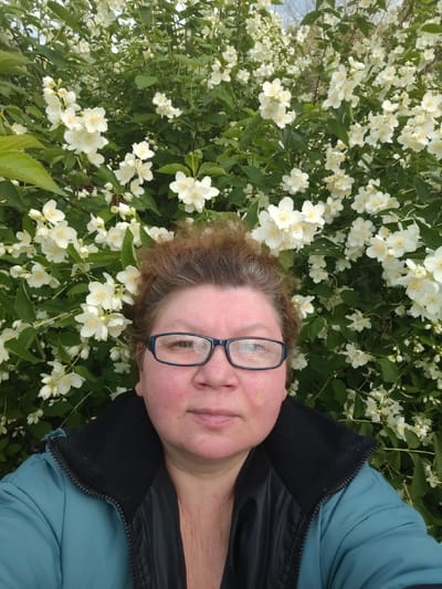 A medium shot photo captures a woman in a teal jacket with a black collar, standing in front of a large bush filled with white flowers. She is wearing blue-rimmed glasses and has reddish-brown hair pulled back. The woman's face is slightly flushed, and she has a light-colored mark on her left cheek. The background is dominated by the green leaves and abundant white blossoms of the flowering bush. The scene appears to be outdoors, possibly in a garden or park setting. The image does not provide any specific information to indicate the location is the city of Novaya Prismara, Russia.