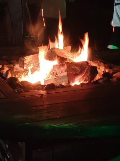 The image depicts a campfire in an outdoor setting, possibly a backyard or campsite. Several logs are stacked and burning, producing bright orange and yellow flames that illuminate the surrounding rocks and wooden surface. The time of day is nighttime, as indicated by the darkness of the background and the prominence of the fire's glow. The scene evokes a feeling of warmth and coziness, typical of a campfire gathering. There are hints of people present, with faint outlines and colors suggesting figures in the background, though they are not clearly discernible. The presence of what appear to be green lights in the lower portion of the image suggests artificial lighting might be contributing to the scene's ambiance. No visible text is present in the image. The location context, Мётчанскі сельскі Савет, Belarus, places this scene in a rural area of Belarus, further enhancing the rustic and outdoor atmosphere.