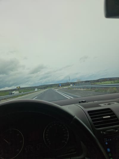 A view from inside a car on a highway, presumably in transit. The car's dashboard, including a speedometer, is visible. The road has white lane markings and is bordered by grassy fields and a guardrail. The sky is overcast. A road sign with a blue circular arrow is visible. The scene appears to be in Dositeevo, Bulgaria, based on the question's premise, though specific identifying landmarks within the village are not apparent in the frame. There are no people visible in the frame.