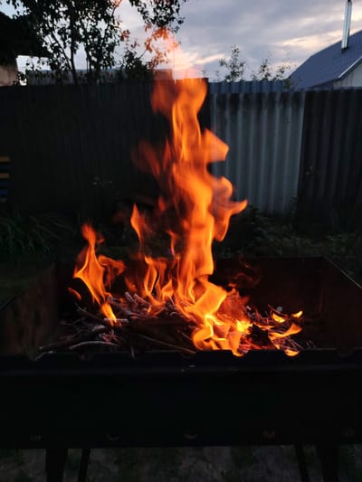 A barbecue pit is ablaze with orange flames and burning wood. In the background, a grey corrugated metal fence runs horizontally across the image. Beyond the fence, the silhouette of trees can be seen against a cloudy sky at dusk. To the right, a portion of a house with a blue roof and a chimney is visible. The scene appears to be outdoors, possibly in a backyard or a rural area. The location is identified as Shklov, Belarus.