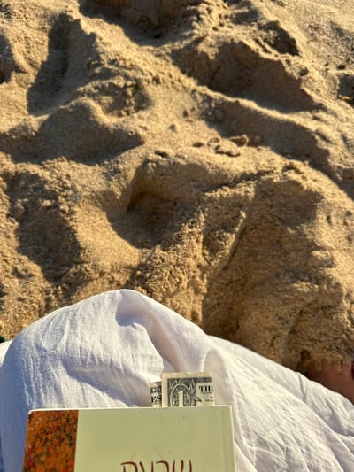 The image depicts a sandy beach scene within the municipality of Mafra, Portugal. The upper two-thirds of the frame consists of light-colored, fine-grained sand, exhibiting natural undulations and shadows consistent with dunes under direct sunlight. In the foreground, a person is partially visible, largely covered by white, creased fabric, likely linen. A book with a light-colored cover and an abstract orange and red design on its visible edge rests on or is held by the fabric. A folded United States one-dollar bill is inserted into the book, functioning as a bookmark, with portions of the "ONE" denomination and the obverse design visible. A small section of skin, likely a foot or lower leg, is minimally discernible on the far right, adjacent to the fabric. The overall scene suggests a person resting or reading on the beach.