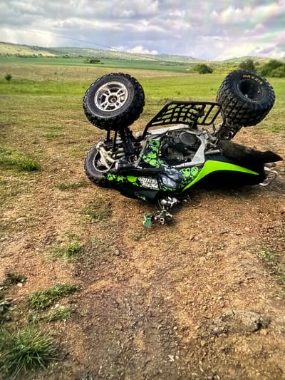 A four-wheeled ATV is on its side in a field. The ATV is primarily silver and black with green accents and skull designs. It is lying on a patch of dirt and short grass. The surrounding area is mostly green grass and farmland, stretching towards a horizon under a cloudy sky. The location is not specified, and there is no information to ascertain this image came from the city of Petarch, Bulgaria.