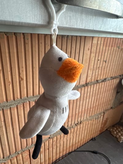 A light gray plush toy bird with an orange beak and black feet hangs from a white plastic hook against a brick wall. The wall is constructed of vertically oriented orange bricks with visible mortar lines. The bird is suspended by a thin string. In the lower right of the frame, a black cable lies on a dark gray surface, and a small, egg-carton-like object is visible. The scene is outdoors, likely in Otradny, Russia.