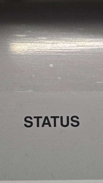 This is a close-up, vertically oriented photograph depicting a light gray, textured surface. The word "STATUS" is printed in uppercase black sans-serif font, horizontally centered in the lower half of the frame. A subtle, faint horizontal crease or line extends across the surface approximately one-third of the way down from the top edge, just above the text. The upper portion of the image shows a gradient, transitioning from a darker gray at the very top to a lighter gray band below it. This lighter upper band exhibits multiple horizontal streaks of bright reflection, indicative of an overhead light source interacting with a semi-glossy or uneven surface texture. Several small, irregular white spots and numerous dark fibrous specks, consistent with dust or lint, are visible across the entire light gray surface. No individuals are present. No actions or interactions are depicted. This scene is situated in Lompoc, United States.