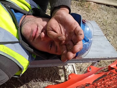 A close-up, over-the-shoulder shot shows a male, possibly a construction worker, lying on his back outdoors in Raygorodok, Kazakhstan. He is wearing a reflective yellow and grey vest over a blue shirt and a dark grey hooded jacket. His face is partially obscured by his dirt-covered hand, which is resting on his forehead. Safety glasses with a blue frame are visible near his head, and an orange plastic safety fence is in the foreground. The ground appears to be a mix of gravel and dirt with sparse vegetation. The lighting suggests it is daytime.