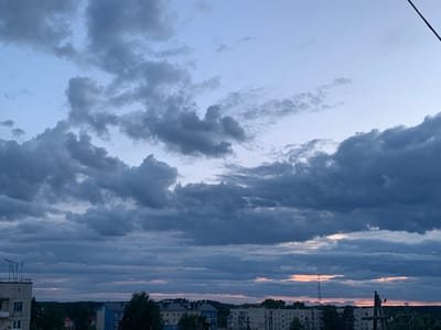 The image depicts a dusky sky over the city of Volchansk, Russia. Dark, heavy clouds dominate the upper portion of the frame, with lighter patches of blue sky peeking through. A strip of pale orange and pink hues suggests the setting sun near the horizon line. Below the sky, the silhouette of a cityscape is visible, with the tops of apartment buildings and trees. On the left side, a concrete building with an antenna on its roof is partially visible. Electrical poles with wires are present in the mid-ground and right foreground. The overall atmosphere is one of a cloudy evening in an urban environment.