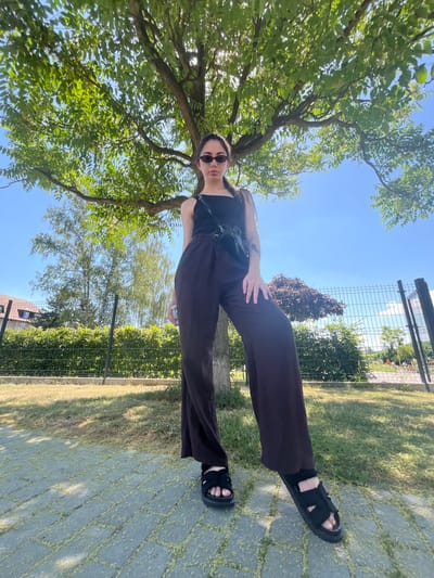 The image shows a young woman posing outdoors. She is wearing dark, wide-leg pants, a black tank top, black sandals, and sunglasses. A black bag is slung across her chest. The woman is standing on a paved walkway that is bordered by a grassy area and a chain-link fence. Behind the fence, there is more greenery and some buildings in the distance. A large tree with lush green leaves is to her left, and the bright blue sky is visible above. The sunlight suggests it is daytime. The scene appears to be a park or a residential area. The woman's posture and gaze suggest she is posing for a photo. The image is shot from a low angle, looking up towards the woman and the sky. The provided location context is Chotěšov, Czechia.