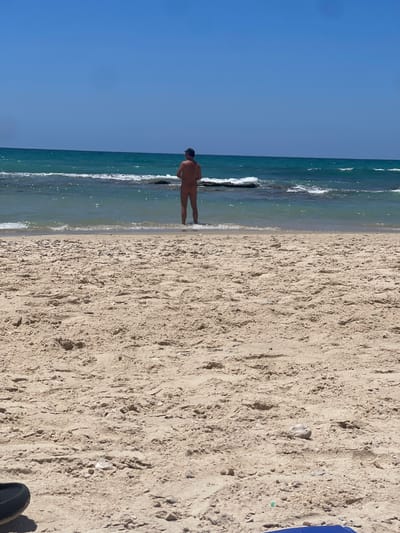 A person is standing in shallow water at the edge of the sea on a sandy beach in Ga'ash, Israel. The person is facing away from the camera, wearing a dark hat and appears to be unclothed. The sea has gentle waves breaking on rocks and is a turquoise color, with a clear blue sky above. The foreground is dominated by tan sand with some small stones and debris.