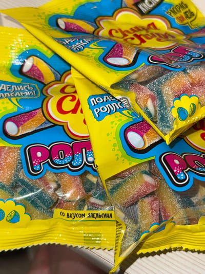 The image shows two bags of Chupa Chups sour rolls, a type of candy. The bags are bright yellow with blue and pink accents and feature cartoonish designs of the candy. The candy itself is visible through the clear plastic packaging, appearing as colorful, sugar-coated rolls with a soft, chewy texture. Text on the packaging is visible in Russian, including "ДЕЛИСЬ ВКУСАМИ!" which translates to "Share the flavors!" and "СО ВКУСОМ АПЕЛЬСИНА" which means "With orange flavor." The bags are placed on a light-colored, textured surface, likely a table. The lighting suggests it is daytime, and the overall impression is of a casual, consumer product display. There are no people or animals visible. The scene appears to be indoors, given the lighting and the surface it is resting on.