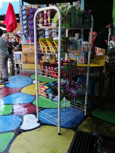 The image captures a scene outside a small shop in Tegalwangi, Indonesia, likely a street vendor or a small convenience store. The ground is a colorful, wet surface painted with large, irregular shapes in shades of blue, red, yellow, and green, suggesting it might be a public space or a painted walkway that has recently rained.
Dominating the foreground is a white, multi-tiered metal display rack filled with various packaged snacks and candies. The snacks are mostly in colorful, crinkly bags. Some bags are a vibrant yellow, others purple, and some have red and white packaging. There are also some boxes of candy, including one with cartoon animals and the text "Bubble Stick".
To the left of the display rack, a person wearing a grey shirt and pants is partially visible, facing away from the camera. Two young boys stand near the shop's entrance. One boy, wearing a dark t-shirt, is looking towards the display rack, while the other, in a lighter brown shirt, is looking at the camera with a surprised expression. Behind them, a bright red plastic bag hangs from a hook.
The shop itself appears to be constructed from wood, with a yellow-painted facade. A glass display case is visible on the left side of the shop entrance. In the background, more packaged goods hang from the ceiling or walls of the shop. A woman in a dark shirt and patterned skirt is partially visible behind the display rack, tending to the shop. A woven basket is placed on a table to her right. The overall impression is of a lively, local marketplace or a small retail establishment in a tropical environment, possibly during or after a rain shower due to the wet ground.