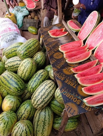 A vibrant outdoor market scene features a large pile of whole watermelons with green striped rinds, juxtaposed against cut watermelon slices displaying their bright red flesh. The slices are neatly arranged on a dark surface marked with repeated logos. A partial bag of animal feed and a crate of pineapples rest nearby. People are visible in the background; one wearing a blue shirt, holding a bag, and another sitting while holding a white bag.