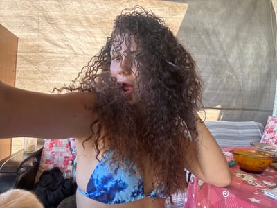 A woman with long, curly brown hair is taking a selfie. She is wearing a blue and white tie-dye bikini top. Her mouth is open, and she appears to be making a playful expression. Her arm is extended towards the camera, as if holding a phone. In the background, there is a table covered with a pink tablecloth with cartoon characters on it. On the table, there are two glass bowls, one of which contains a yellow-orange soup or stew. There are also pillows on a sofa in the background. The overall environment suggests a casual outdoor setting, possibly a backyard or patio. The image does not contain any information that would identify the location as Belopoptsi, Bulgaria.