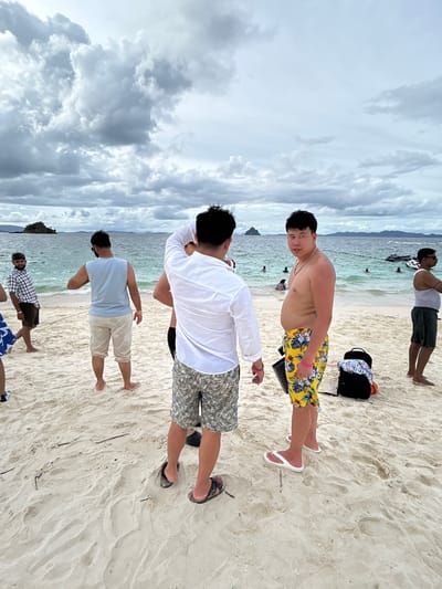The media file shows a group of people on a sandy beach in Tambon Phru Nai, Thailand. The scene is set with the ocean in the background, the water appearing calm and the sky filled with dramatic, cloudy formations. The overall atmosphere is that of a tropical beach day, though the weather appears overcast.
Several individuals are visible, mostly men, dressed in casual beach attire. One man in the foreground is shirtless, wearing patterned swim trunks, and appears to be looking at the camera. Another man, facing away from the camera, is wearing a white long-sleeved shirt and patterned shorts. Further back, a man in a light blue t-shirt and light-colored shorts is also visible. Another person, at the very top of the frame, is wearing a white tank top and dark shorts. Beach bags and some personal items are scattered on the sand.
The people appear to be socializing and enjoying their time at the beach. The cloudy sky suggests it might be later in the day or that the weather is changeable. No text is visible in the media file.