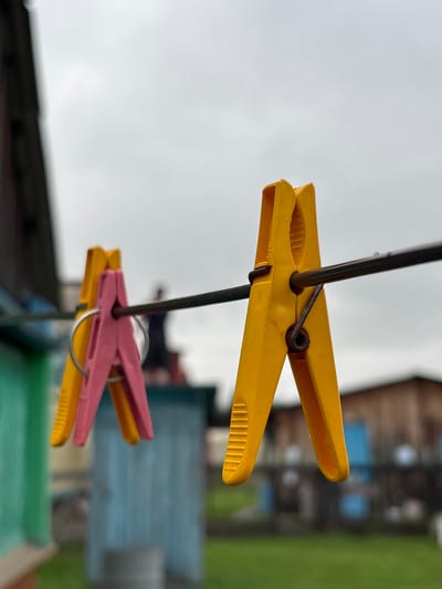 In Gonja, Russia, under a cloudy sky, two clothespins, one pink and one yellow, are clipped to a clothesline. The pink clothespin is on the left, and the yellow one is to its right, slightly overlapping. Behind them, a blurry figure stands near a light blue structure. To the left, a green structure is visible, and in the background, several indistinct wooden buildings stand amidst greenery. The focus is on the clothespins, with the background rendered out of focus.
