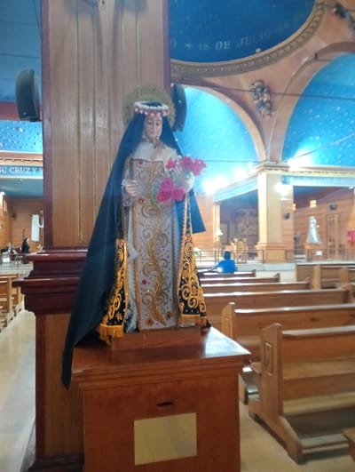 A statue of a saint with flowers stands on a wooden platform inside a church. The saint is dressed in a white and gold robe with a black shawl and a flower crown. Rows of wooden pews are visible in the foreground and background. A person in a blue shirt sits in one of the pews. The church has wooden columns, arches, and a blue ceiling with painted stars. "Pozo Almonte, Chile" is inferred as the location based on visual context of similar religious images in the area.