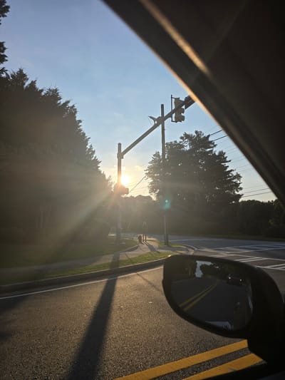 This image depicts a view from a vehicle on a sunny day, likely in Marietta, United States. The sun is low in the sky, casting long shadows and creating a lens flare effect. In the foreground, the asphalt road with yellow lane markings is visible, along with a concrete curb and a sidewalk. A person and a dog are walking on the sidewalk, with trees and foliage lining the background. A traffic light is positioned over the intersection. The side mirror of the car reflects the road behind, showing a red vehicle. The scene suggests a suburban environment with a clear blue sky.