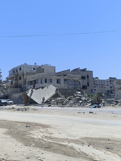 The image depicts a street scene in Gaza, showing heavily damaged buildings under a clear blue sky. The structures are in a state of ruin, with debris scattered across the ground. Several cars are parked on the left side of the road. The foreground is a sandy, unpaved area with tire tracks and scattered debris. The overall impression is one of devastation.