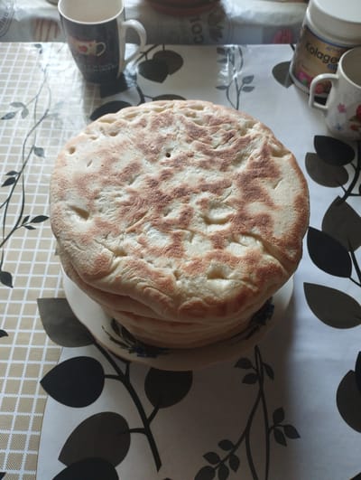 In the city of Ciuciulea, Moldova, a stack of flatbreads sits on a plate, which rests on a table covered with a patterned tablecloth. The flatbreads have a lightly browned and textured surface, with small indentations. To the left of the flatbreads, a mug with a coffee cup design is visible. To the right of the flatbreads, another mug and a white jar labeled "Kolage ACTIVE" are present. The tablecloth features a checkered pattern in the upper left corner and a floral or leafy design in the rest of the visible area. The lighting suggests it is daytime, possibly indoors.