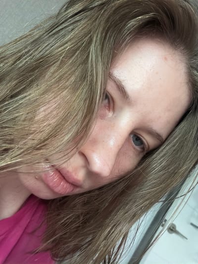 A close-up, angled shot shows the face of a young woman with light brown hair. Her hair is draped across her face, obscuring part of her forehead and eye. She is wearing a bright pink top. The background appears to be a neutral, light-colored wall, with a hint of what might be a door or cabinet on the right side. The lighting is soft and natural. No specific location in Dongducheon-si, South Korea can be determined from the image.