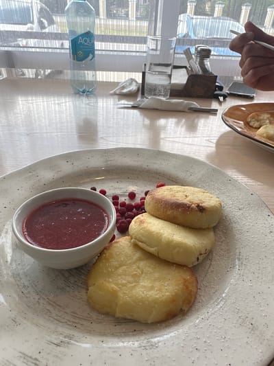 In Slyudyanka, Russia, a close-up shot captures a breakfast setting on a wooden table. In the foreground, a white plate holds three golden-brown syrniki (cheese pancakes) dusted with powdered sugar. To the left of the syrniki, a small white bowl contains a vibrant red berry sauce. Scattered around the syrniki and sauce are fresh red berries, likely lingonberries or cranberries.
In the background, a clear bottle of "Aqua Mineral Water" stands prominently. To its right, a glass filled with a fizzy drink and ice sits next to a napkin dispenser containing utensils and salt and pepper shakers. Further in the background, a person's hand holds a fork, indicating they are about to eat. The scene is illuminated by natural light filtering through windows, casting reflections on the polished wooden surface of the table. The overall atmosphere suggests a relaxed meal in a cafe or restaurant.