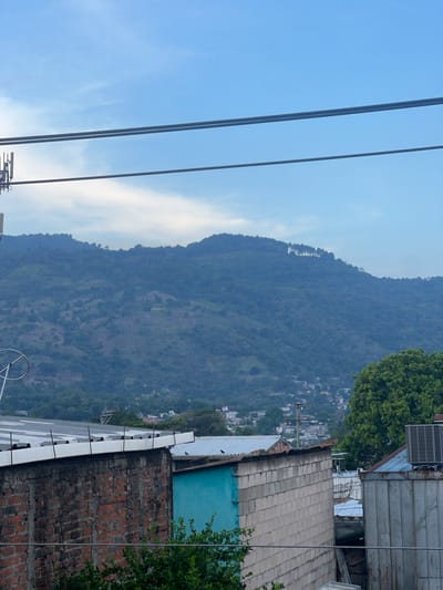 The image shows a mountain range viewed from an elevated position in San Salvador, El Salvador. Several utility lines cross the left portion of the frame against a blue sky with light clouds. Buildings and trees are visible on the right. A brick wall and roofline occupy the lower right.