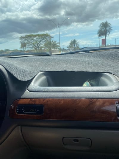 The image, taken from inside a vehicle and rotated 90 degrees clockwise, presents a bright daytime view through the windshield. The interior of the car features a textured grey fabric dashboard cover on the left and a polished, dark brown wood-grain trim on the right. A dark grey plastic air vent is prominent in the middle of the dashboard, with a small, reflective object visible inside it.

On the wood-grain panel, a rectangular digital clock displays "2:01", likely indicating the time, with small "H", "M", and "S" buttons below it.

Outside the vehicle, a partly cloudy sky with clear blue patches is visible. The landscape is dominated by lush green trees, including several tall palm trees characteristic of a tropical or subtropical region. Utility poles or streetlights line the roadside, and a faint red rectangular structure can be seen in the distance. The bright conditions and the nature of the vegetation are consistent with an outdoor scene in La Cruz del Pastel, Venezuela, during the early afternoon, matching the 2:01 time on the display.