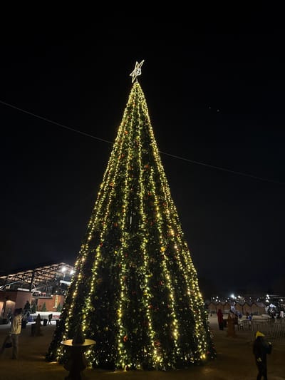 A brightly lit Christmas tree dominates the dark night, adorned with strings of lights and red ornaments. A radiant star tops the tree. Below, a structure appears to be a building or stage, with people milling around. The ground is visible, suggesting an outdoor setting. A few overhead lights illuminate the background, adding a soft glow to the scene.