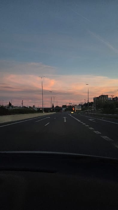 The image displays a multi-lane asphalt road in Paterna, Spain, viewed from within a vehicle during crepuscular hours. The sky transitions from deep blue overhead to a band of pink and orange stratocumulus clouds near the horizon, with faint contrails present. The vehicle's dark dashboard occupies the bottom portion of the frame. The road features white solid and dashed lane markings, including a prominent straight-ahead arrow in the center lane and a left-turn arrow in the adjacent lane. Numerous tall, dark-colored streetlights line both sides of the roadway, extending into the distance. On the right side, a traffic signal displays an amber light. Adjacent to this, a rectangular white informational sign is visible, followed by a circular speed limit sign indicating "80" kilometers per hour. Buildings, consistent with an urban or semi-urban environment, are present on the right horizon line, interspersed with vegetation. A concrete median barrier runs along the left side of the carriageway, and an overhead bridge structure is discernible further down the road. No human subjects are depicted.