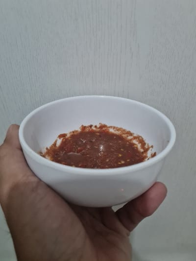 A person holds a small white bowl containing a reddish-brown sauce. The sauce appears to have small particles and possibly some red pepper flakes visible within it. The bowl is held tilted slightly, exposing most of the sauce. The background appears to be a light-colored surface with subtle texture.