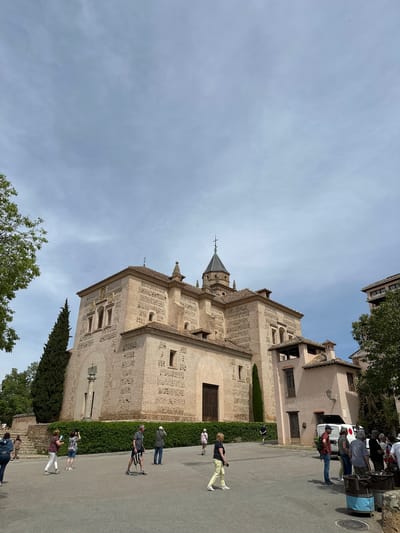 The image captures a large, historic building, likely a church or monastery, under a clear to partly cloudy daytime sky in Granada, Spain. The building features intricate brick and stone patterns on its facade, including decorative geometric designs, multiple arched windows, and a prominent central steeple topped with a cross. Its roof is tiled, and sections of the exterior show lighter-colored stone contrasting with the brickwork.
Along a paved road or pathway to the right of the building, several people are visible. Some individuals are walking, seemingly sightseeing, while others are gathered in groups. One person in light yellow pants and a patterned top walks towards the foreground, possibly looking at a phone. Further back, a group of people stands near a white van parked on the side of the road, suggesting a tour group or a stop. Lush green hedges line the base of the building, and scattered trees with green foliage are present in the foreground and background. The overall scene depicts a sunny day with visitors exploring a historic architectural site. No visible text is discernible in the image.