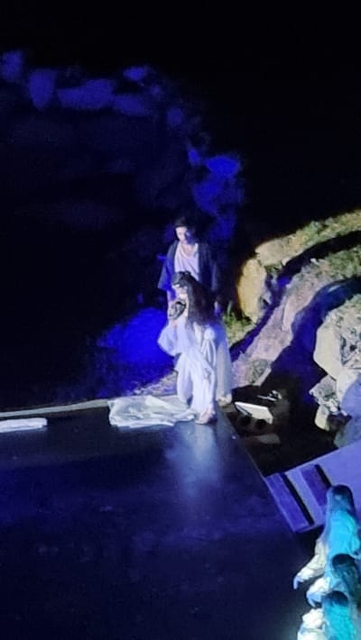 In the city of Sant Joan les Fonts, Spain, two figures are illuminated on a darkened stage. The foreground is a dark, reflective surface, likely a stage floor, with subtle blue lighting. To the right, a rocky incline is visible, also bathed in dim light.

The main subjects are two people standing on a raised platform near the rocky incline. The person in front is a woman dressed in a flowing white gown with elaborate sleeves, her long dark hair partially obscuring her face. She appears to be kneeling or in a low crouch, holding a white fabric on the stage. Behind her stands a person in a dark robe over a lighter-colored garment, with dark hair. They are looking down towards the woman.

Further to the right and in the lower part of the image, two more figures are visible. The topmost figure is wearing a light-colored garment and appears to be holding something. Below them, another figure, also in a light-colored garment, is partially visible. The overall scene suggests a theatrical performance, possibly a dramatic or interpretive dance, with a focus on ethereal or spiritual themes suggested by the lighting and costumes. The environment is dark, with strategically placed lighting creating pools of illumination and deep shadows.