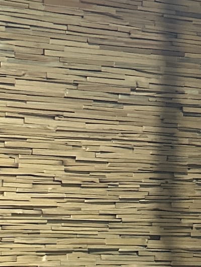 The image displays a close-up, abstract view of a textured surface composed of numerous vertical, thin wooden elements. These elements appear to be reeds or thin strips of wood, closely packed together, creating a sense of a wall or screen. The lighting casts subtle shadows, highlighting the depth and irregularities of the surface. The color palette is dominated by natural wood tones, ranging from light beige to muted browns, with some darker areas suggesting shadows or natural variations in the material. There are no discernible subjects like people or animals, nor is there any visible text. The overall impression is one of a natural, perhaps rustic, decorative material. The context provided, Prokhladny, Russia, does not offer specific visual cues within the image itself, so the description focuses solely on the visual elements present.