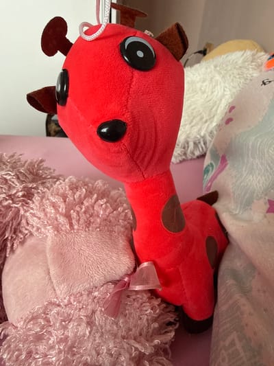 The media file is a close-up, top-down shot of several plush toys arranged together. The primary subject is a red ladybug-shaped plush toy with black spots. It has large, black button eyes with white highlights and small brown antennae and legs. A thin white cord is attached to its head. To the right and partially behind the ladybug is a fluffy pink plush toy, possibly a sheep or llama, adorned with a small pink bow. In the background, other plush toys are visible, including one with a white furry texture and another with a patterned fabric featuring waves and heart shapes, possibly a unicorn or cloud-themed item. The toys appear to be on a soft surface, possibly a bed or couch, given the presence of fabric and the casual arrangement. The lighting suggests an indoor setting, likely during the daytime. There are no visible people or text in the image. The context provided indicates the location is Rogosh, Bulgaria.