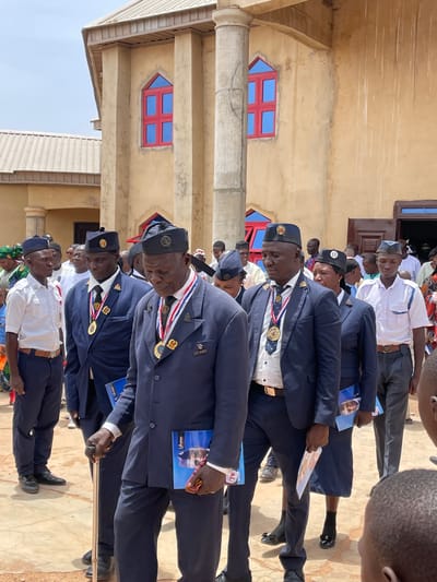 The image depicts a formal outdoor gathering of men in Ladura Foron, Nigeria, standing in front of a light brown building under bright daylight. Most of the men, predominantly older, are dressed in dark blue uniforms featuring suits, white shirts, ties, and hats, some adorned with medals and sashes in red, white, and blue. Other men wear white short-sleeved shirts, dark trousers, and caps. Their expressions are largely serious or contemplative.
Many attendees are holding small pamphlets or programs. Text visible on these includes "Global" and "Gideon" on one, which also features a photo of a man in sunglasses and a mask. Another program shows "TL" or "7L." At least two older men are using walking sticks. The building in the background has distinctive red-framed windows with blue panes, one with an arrow-like design, and features columns and cornices. The ground is a light-colored, possibly paved surface, with some dry earth visible. The clear, sunny weather suggests the event is taking place during the day. In the background, other individuals, some in traditional attire, are present, and a child's head is visible on the far right. The overall scene suggests an official ceremony, parade, or significant communal event.