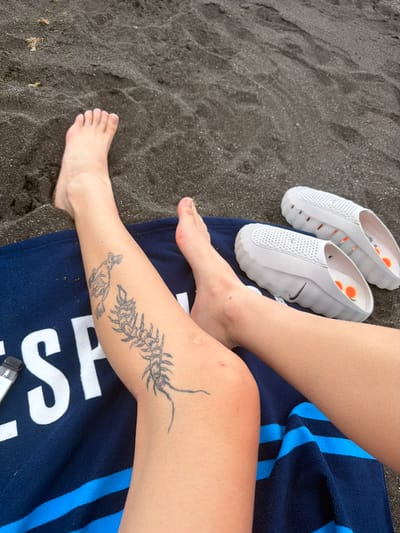 The image shows a close-up view of a person's legs and feet resting on a dark sand beach, possibly in Puerto de la Cruz, Spain. The person is lying on a dark blue towel with blue stripes. On the left leg, there are two tattoos: one of a creature resembling a lion, and another of a centipede or similar segmented insect. Next to the left leg, a small, light-colored object, possibly a vape device, is visible on the towel. A pair of white slip-on shoes with orange accents and a Nike logo are placed on the sand to the right of the legs. The sand is dark and appears to be volcanic, consistent with beaches in Tenerife. The lighting suggests it is daytime.