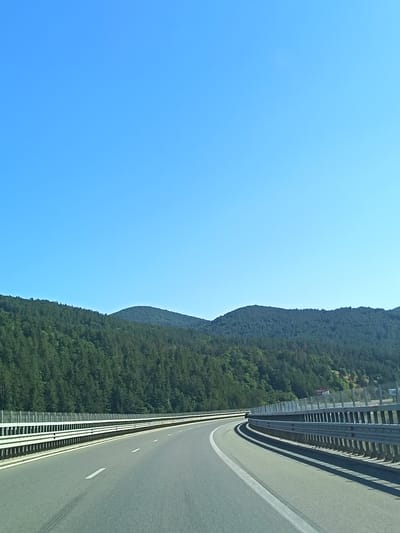 The image depicts a clear blue sky above a winding highway bordered by metal guardrails. The highway recedes into the distance, curving to the left. On the right side of the highway, a dense forest covers rolling hills, with a peak visible in the distance. The overall scene is one of a natural landscape encountered on a road trip. The image does not provide any specific indicators or landmarks that would allow for pinpointing its location within the city of Potop, Bulgaria.